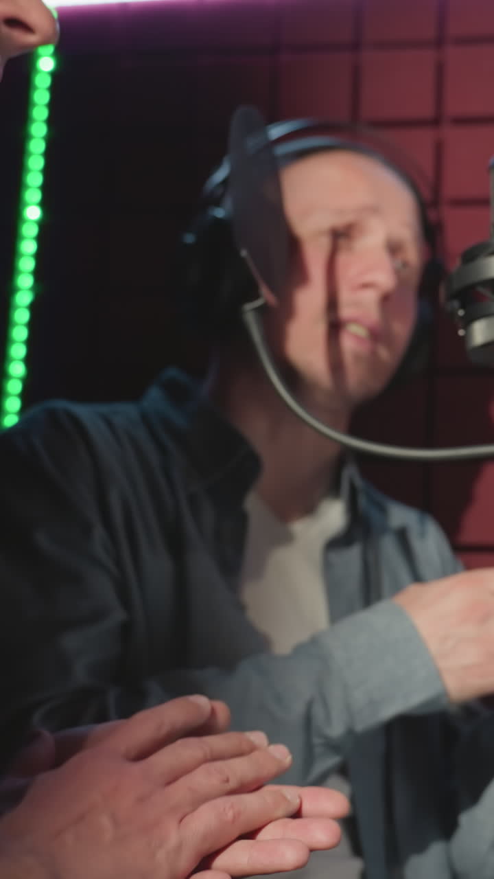 primer plano de dos personas en un estudio de grabación a prueba de sonido, en medio de la actuación con las manos aplaudiendo expresivamente. uno lleva una camisa azul y una camiseta blanca, mientras cantan apasionadamente en un micrófono