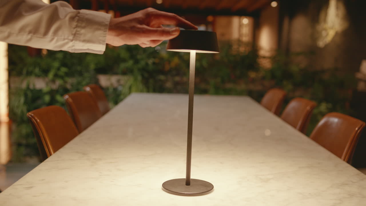 A person turns on a cordless LED table lamp placed in the center of a long table, surrounded by chairs and plants in the background