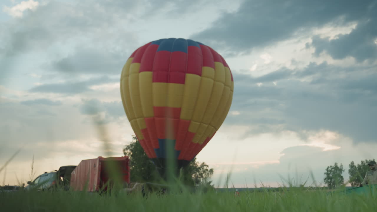closeup of tall green grass blades with man adjusting hot air balloon engine fan while woman jumps excitedly nearby against dramatic cloudy dusk sky over expansive grassy field