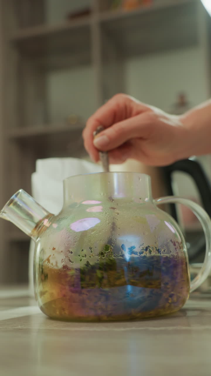 Mano che mescola il tè nella teiera, primo piano del cucchiaio che mescola foglie di erbe in infusore di vetro trasparente, liquido ambrato caldo e vapore delicato, atmosfera da barista artigianale, rituale sul bancone della cucina che enfatizza