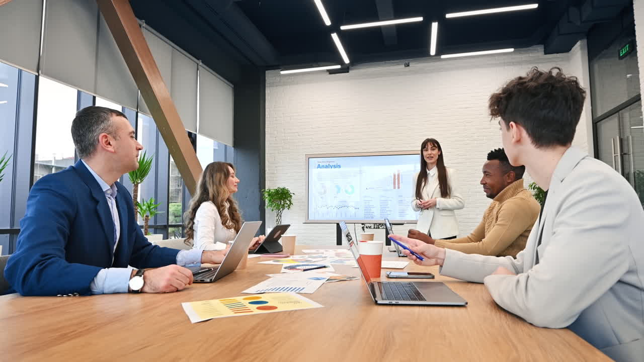 Líder de equipe feminina em reunião de negócios em um escritório, discutindo assuntos de negócios com funcionários usando um display grande com gráficos, documentos e dispositivos eletrônicos na mesa