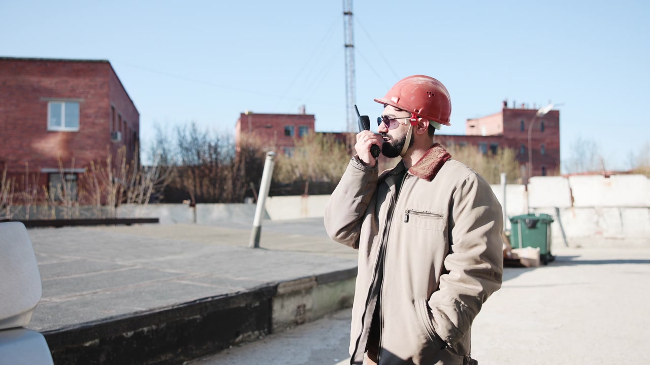 Caposquadra barbuto sul tetto che parla alla radio, indossa casco rosso e occhiali da sole, cappotto pesante, ispeziona i mattoni.