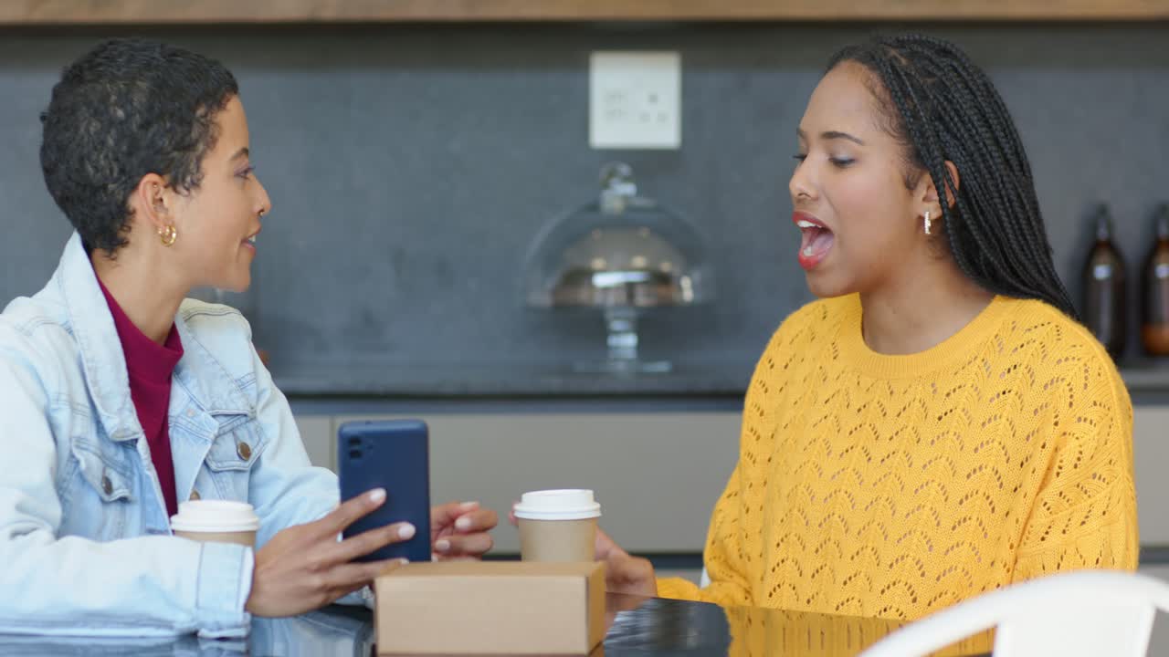 Diverse female friends sitting at cafe showing smartphone and reacting with surprise over coffee