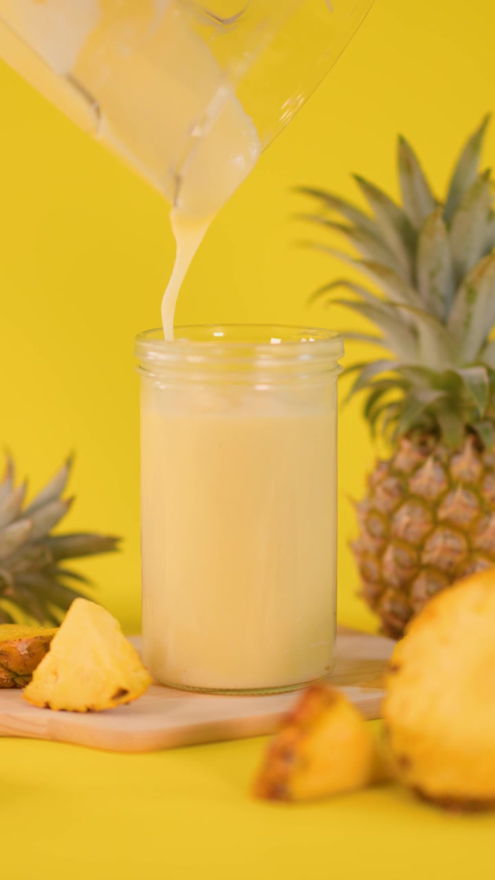 Frullato cremoso all'ananas versato in un bicchiere, circondato da frutta fresca, su sfondo giallo brillante e con illuminazione soffusa.