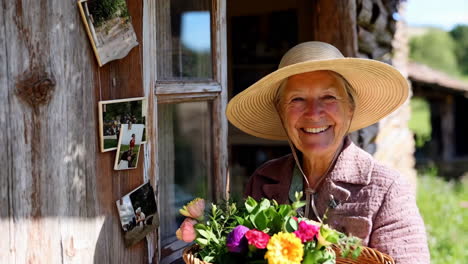 smiling elderly woman in straw hat holding a basket of flowers outside a rustic home