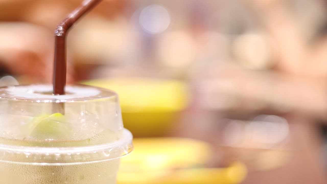 A hand reaches for a plastic cup of iced beverage with a straw on a table in a brightly lit, bustling café environment. Shallow depth of field