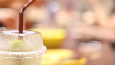 a hand reaches for a plastic cup of iced beverage with a straw on a table in a brightly lit, bustling café environment. shallow depth of field