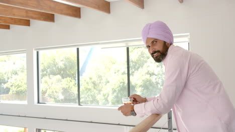 smiling man in turban holding cup of tea, standing by window at home