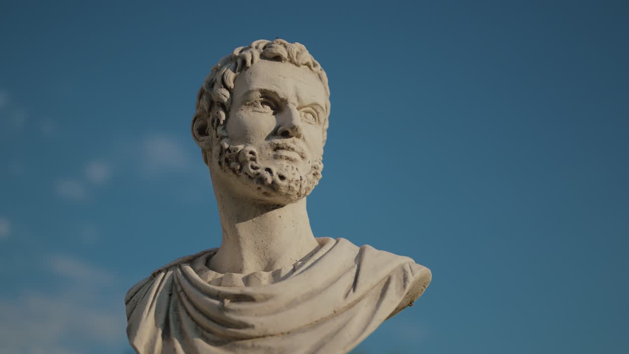Close-up of a classical marble bust statue of a bearded man with detailed facial features and draped clothing against a clear blue sky