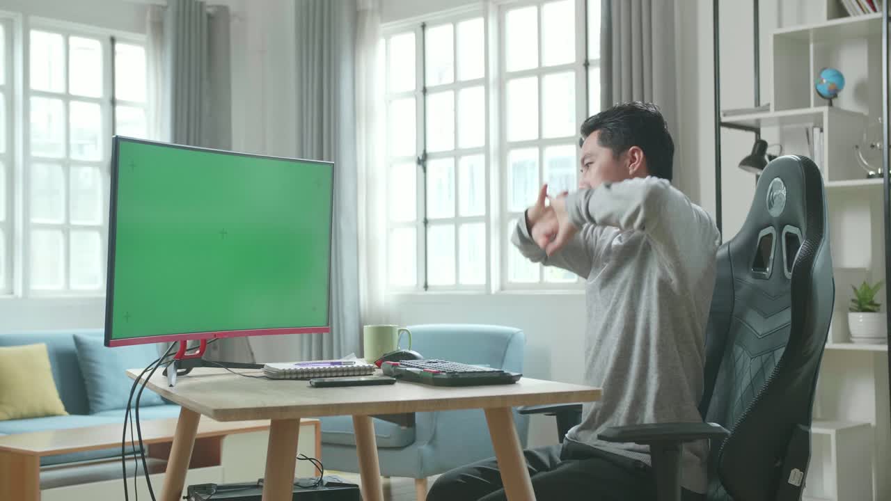 Asian Man In Long Sleeved T-Shirt And Black Pants With Green Screen Desktop Computer Stretching And Leaning Back While Working At Home.