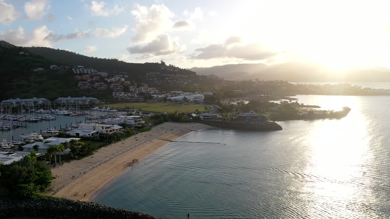 Epic drone shot over Airlie Beach, Queensland as sun sets in background - showing vast marina area beach and with mountain range in background, yachts and boats on sunny evening.