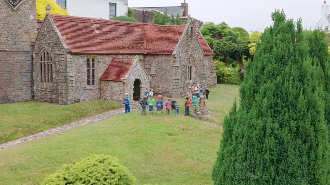 Miniatyr av All Saints Church, Godshill på Isle of Wight mens kameraet panorerer mot venstre over detaljerte modellbryllupsfigurer av brud, brudgom og gjester utenfor steinkirken.
