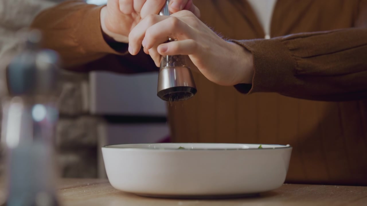 Primo piano di una persona con abiti marroni che condisce il cibo con il pepe in una ciotola bianca su un tavolo di legno. Macinapepe e saliera visibili; lo sfondo include un muro di pietra.