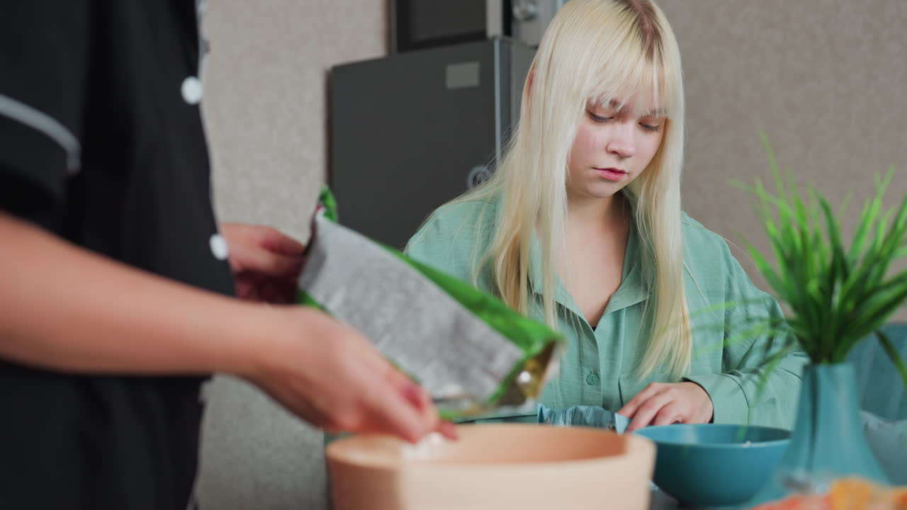 friends in kitchen setting as one girl pours corn flakes from bag into bowl, casual moment, young women preparing snack together, cozy home with modern appliances and relaxed atmosphere