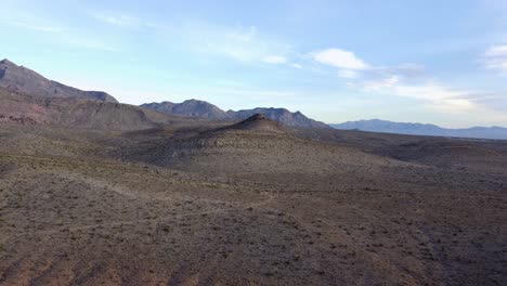 Aerial-shot-of-a-single,-lone-hill-peaking-through-the-desert-landscape-with-stunning-mountains-in-the-background