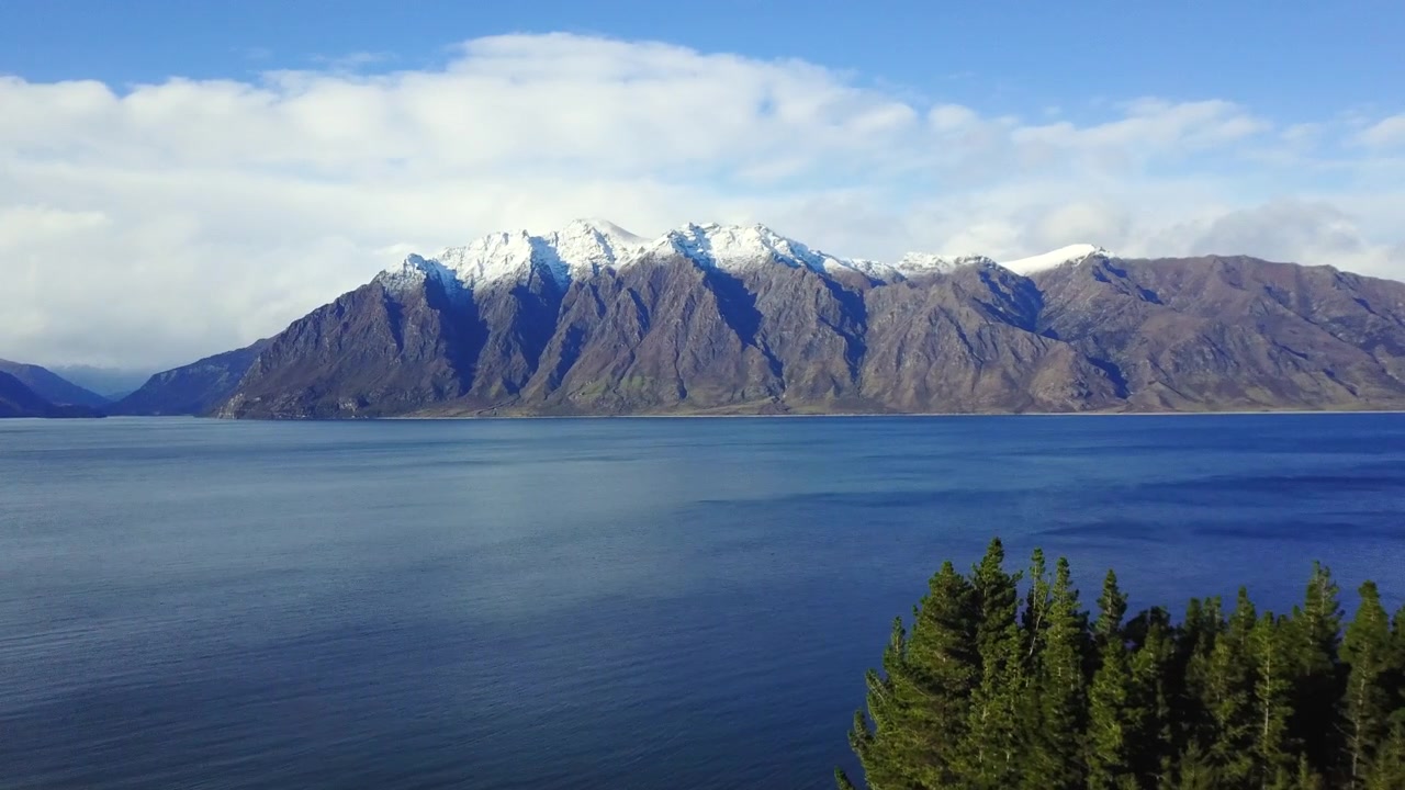 Video aereo di un furgone che guida su una strada costiera con pascoli verdi, poi panoramica sull'oceano blu e montagne innevate