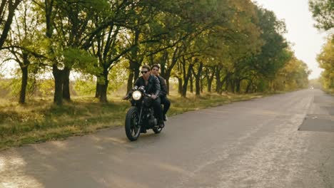 Tracking-shot-of-young-cool-couple-riding-motorcycle-on-forest-road-in-autumn.-Attractive-young-man-in-sunglasses-driving-his