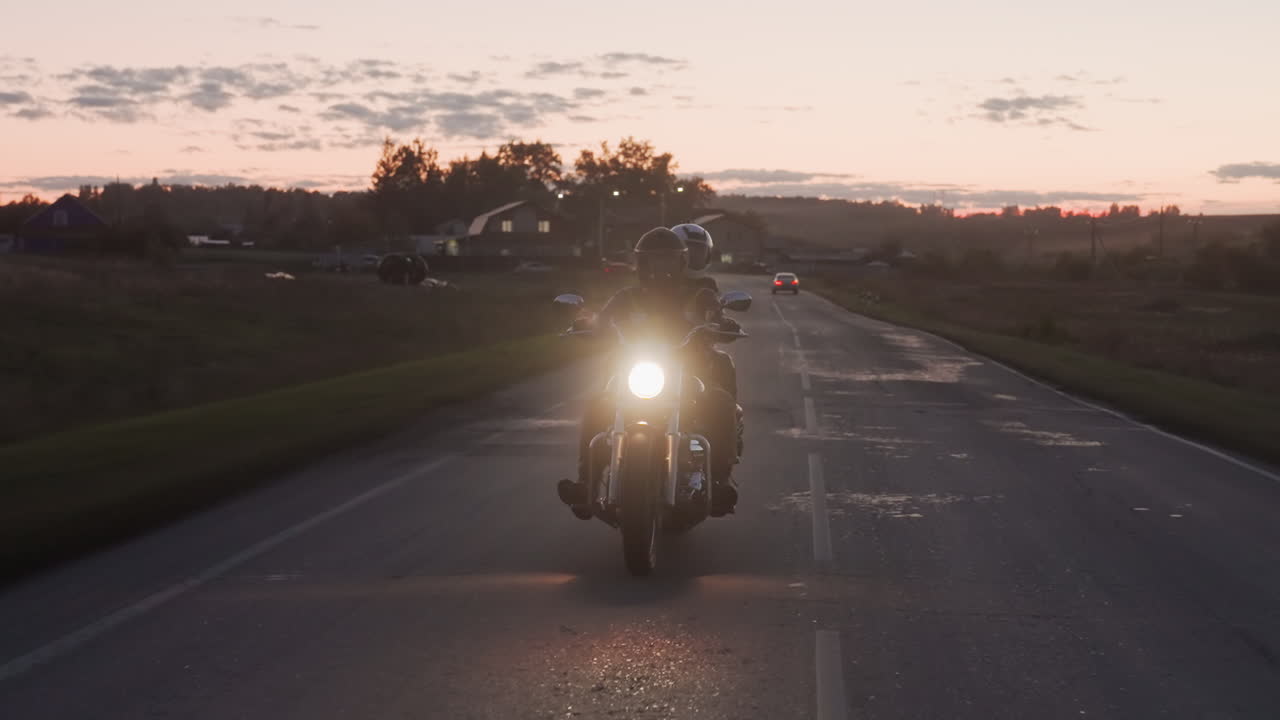 Motorcycle rider with passenger cruises sunset highway, headlamp blazing toward camera, car passes opposite lane, rural horizon glowing with clouds, lens flare and long road lines