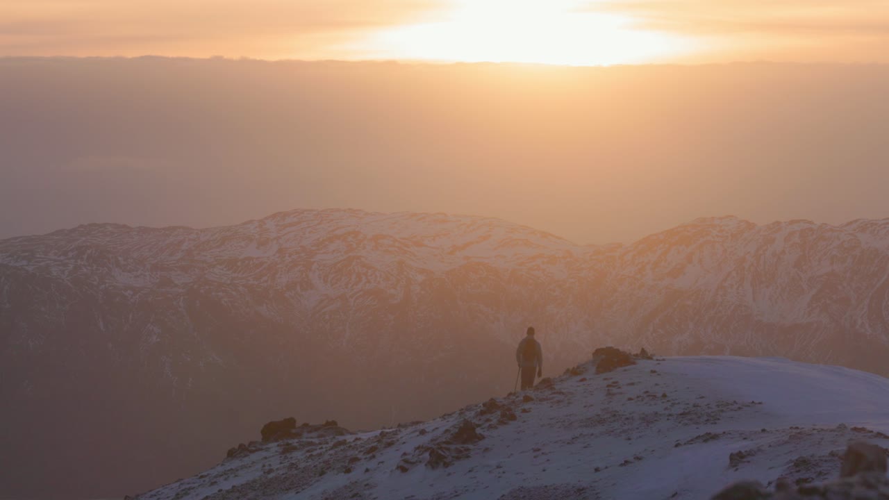 Inquadratura cinematografica di una figura solitaria in piedi su una cresta montuosa innevata durante un magico tramonto all'ora d'oro. Una calda luce arancione illumina le catene montuose a strati, creando una drammatica foschia atmosferica.