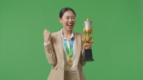 asian business woman in a suit with a gold medal looking at a gold trophy then celebrating winning as the first winner on green screen background in the studio