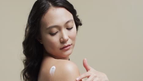 asian woman with dark hair applying cream on her skin on beige background, copy space, slow motion