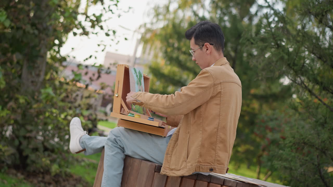 man painting on park bench with portable easel and palette, focused on sketching leafy urban scene near trees wearing brown jacket, jeans and white sneakers late afternoon sunlight casts warm tones