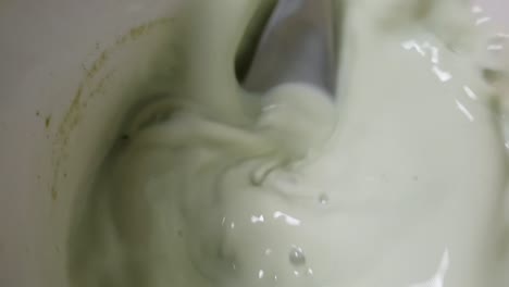 Taza-De-Té-Matcha-Verde-En-Leche-Que-Se-Agita-Para-Beber-Con-Una-Cuchara-De-Acero-Inoxidable-En-Remolinos,-Ondas-Y-Burbujas-A-Cámara-Lenta