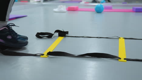 close-up leg view of person in black canvas jumping across agility ladder on gym floor, training equipment, fitness mats, and workout accessories in background