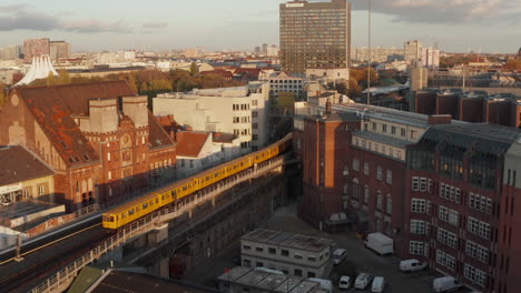 Typischer-Berliner-Gelber-U-Bahn-Zug-Auf-Einer-Anhöhe,-Der-Durch-Das-Stadtviertel-Fährt,-In-Wunderschönem-Sonnenuntergangslicht-Zur-Goldenen-Stunde,-Luftverfolgung-Folgt-Einem-Weitwinkel