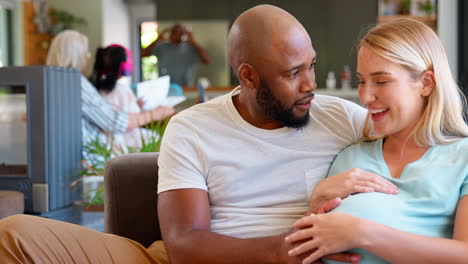 multi-racial couple with pregnant woman on sofa at home with multi-generation family in background