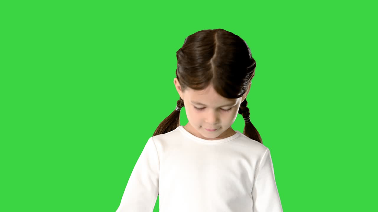 tímida niña con colas de cerdo vestida con una camiseta blanca de manga larga caminando y mirando hacia abajo a sus pies y la cámara en una pantalla verde, clave de croma