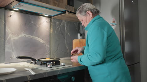 older woman seasoning eggs while cooking them in frying pan on stove in modern kitchen, focused on meal preparation, with kitchen tools, stovetop, and food ingredients visible