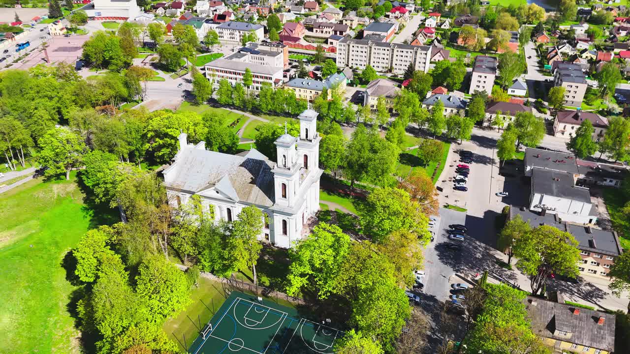 A white church tower stands beside urban basketball courts and leafy trees in the heart of a European city, framed by rooftops and springtime greenery.