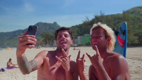 two friends taking happy selfies on a sunny beach with a surfboard