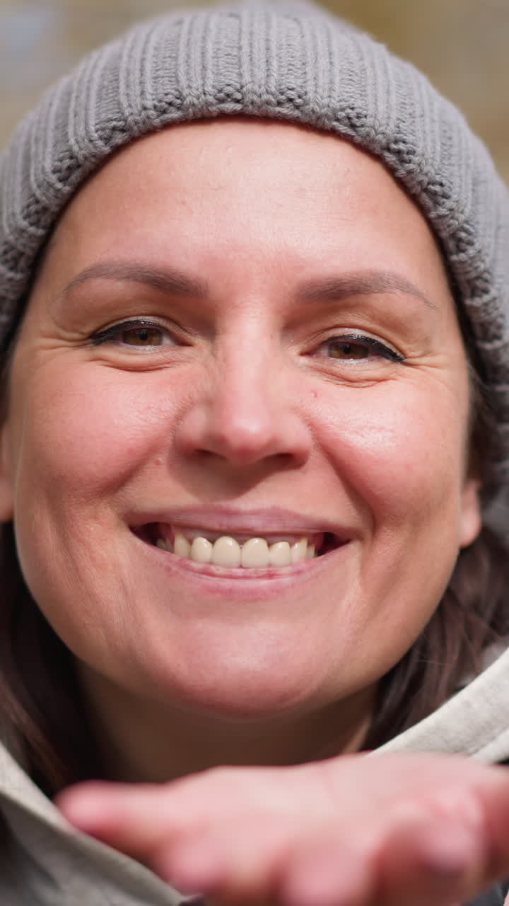 dama alegre con gorro de ceniza y chaqueta rosa soplando hojas secas de su mano con una cálida sonrisa uñas pintadas de negro, rodeadas de árboles borrosos de otoño