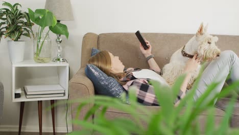 mujer caucásica usando un teléfono inteligente mientras está acostada en el sofá con su perro en casa
