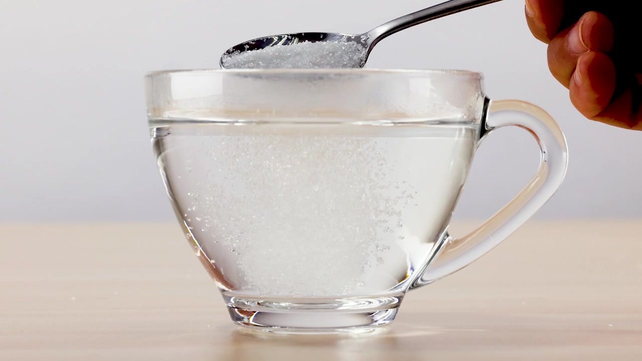 Un cucchiaino di metallo versa zucchero bianco granulato in un bicchiere d'acqua, sciogliendosi gradualmente