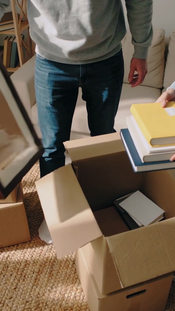 Vertical video: Opening boxes, couple in sweaters moving frame and books in living room unpacking
