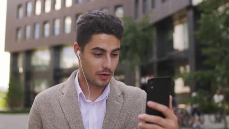 Young-Businessman-With-Earphones-Looking-At-Smartphone-Screen-During-Communication-In-Urban-Environment