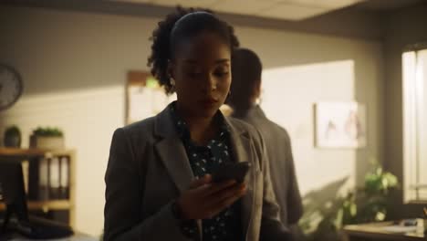 a professional woman wearing a blazer interacts with her smartphone in a well-lit office. she appears focused and engaged in her work while surrounded by an organized workspace.