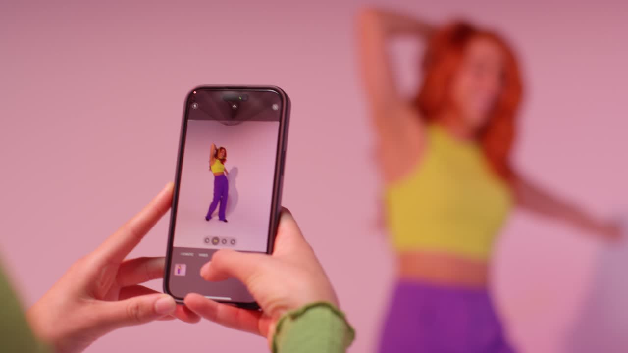 fotografía de estudio de una mujer tomando una foto de su amiga bailando en un teléfono móvil contra un fondo rosa 4