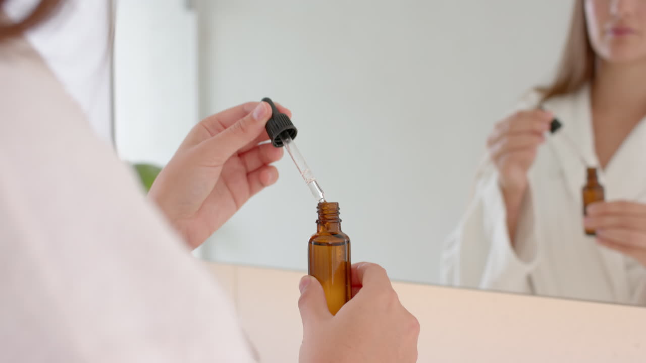 Using dropper bottle, woman applying skincare serum in bathroom mirror reflection, at home