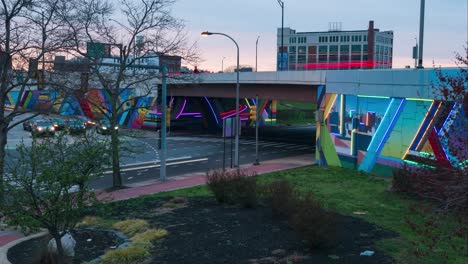 day to dusk timelapse of urban art mural on underpass lights up at sunset in the city car lights pass by on expressway next to park