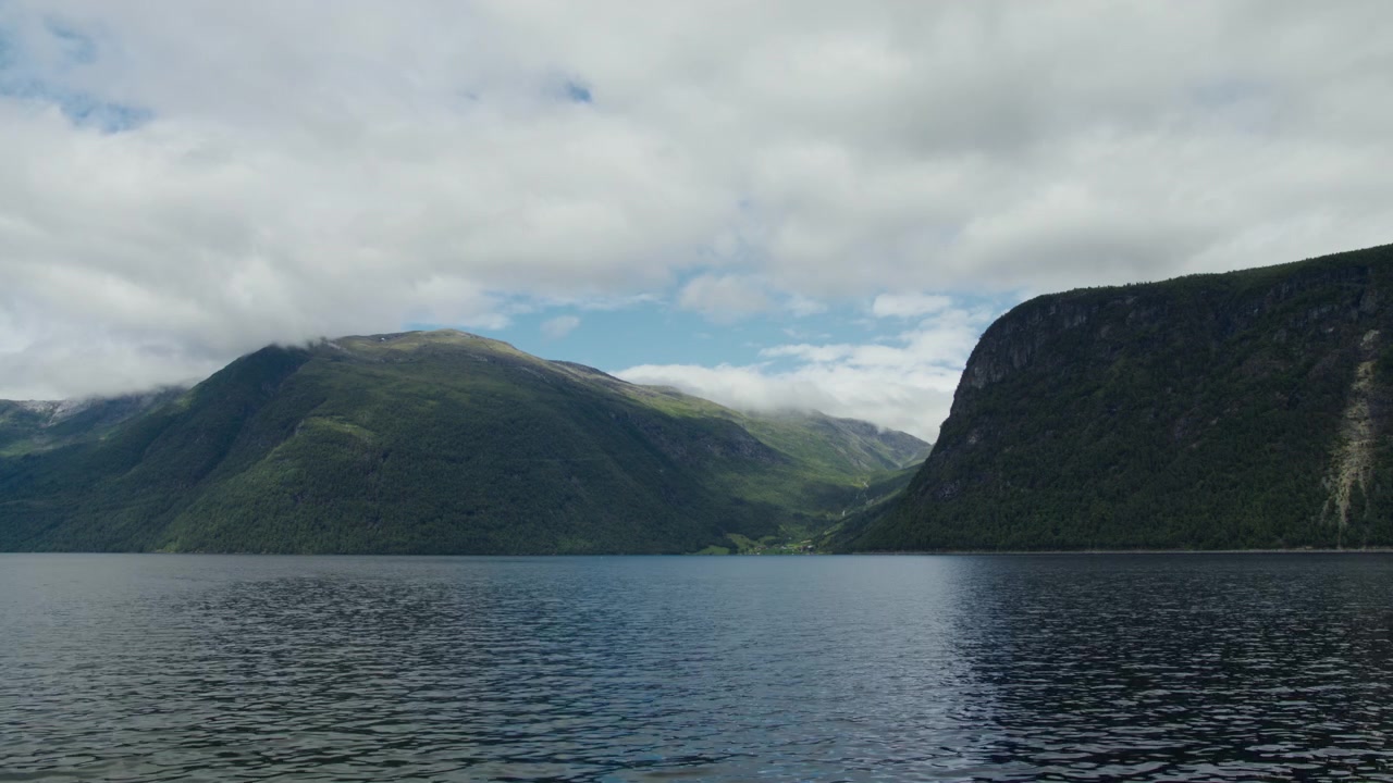 Vista dal basso della telecamera che guarda attraverso un tranquillo fiordo di Sunnmøre verso ripide scogliere boscose sotto un cielo luminoso e coperto