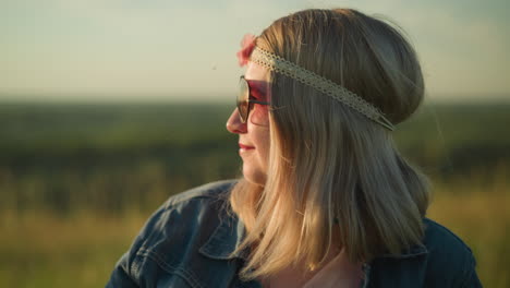 a woman wearing a denim jacket and sunglasses adjusts her hair while smiling in an open field, she has a headband on