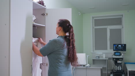 in well lighted clinical room woman therapist retrieves work attire from locker and walks toward x-ray machine adjusting uniform confidently surrounded by medical equipment desk and monitor
