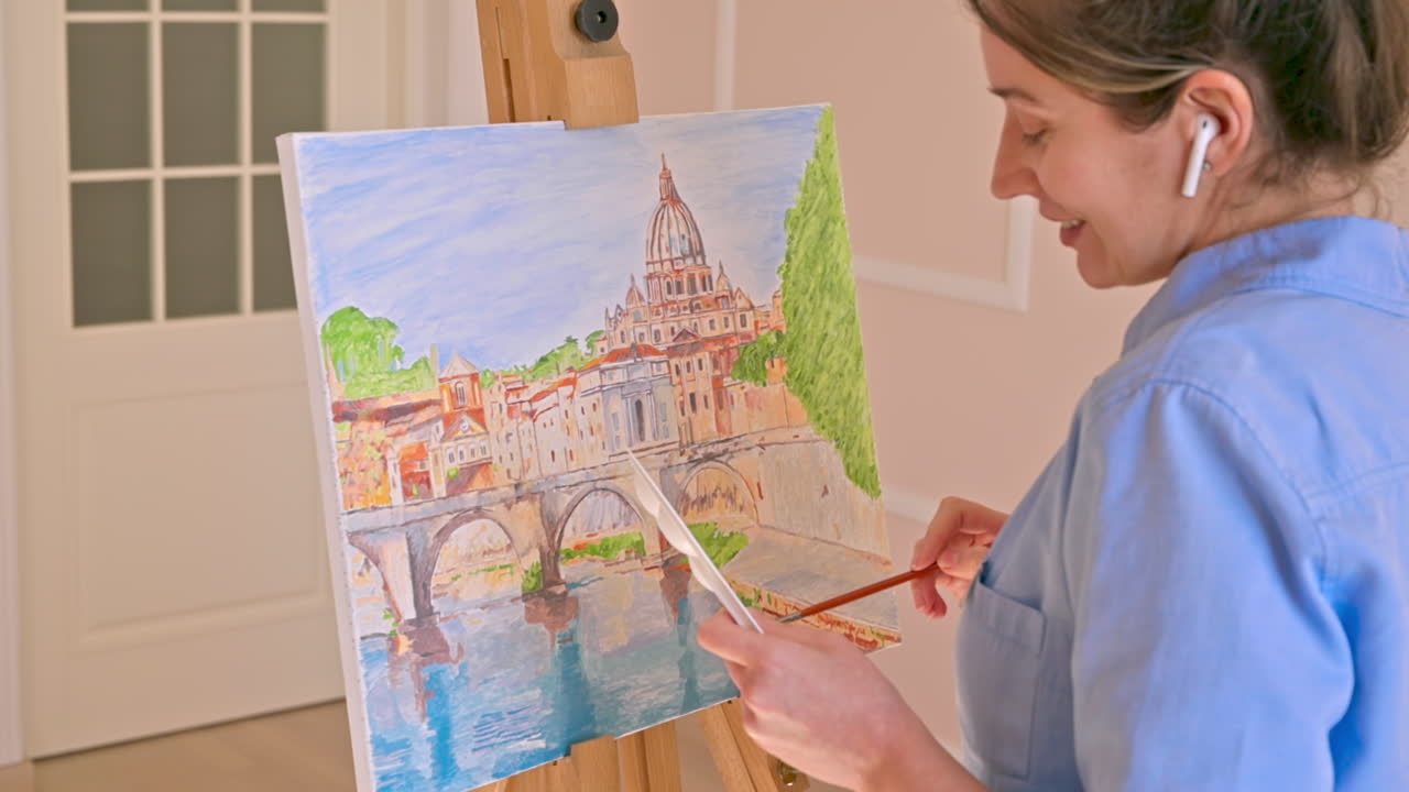 Caucasian woman painting a picture with a palette in her hands and listening music with earphones indoors. Slow motion