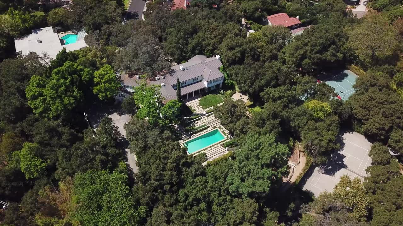 Bird’s eye aerial tracking pass across an upscale Pasadena neighborhood from a top down angle. The shot glides over leafy properties with swimming pools and tennis courts, dense tree canopy, driveways