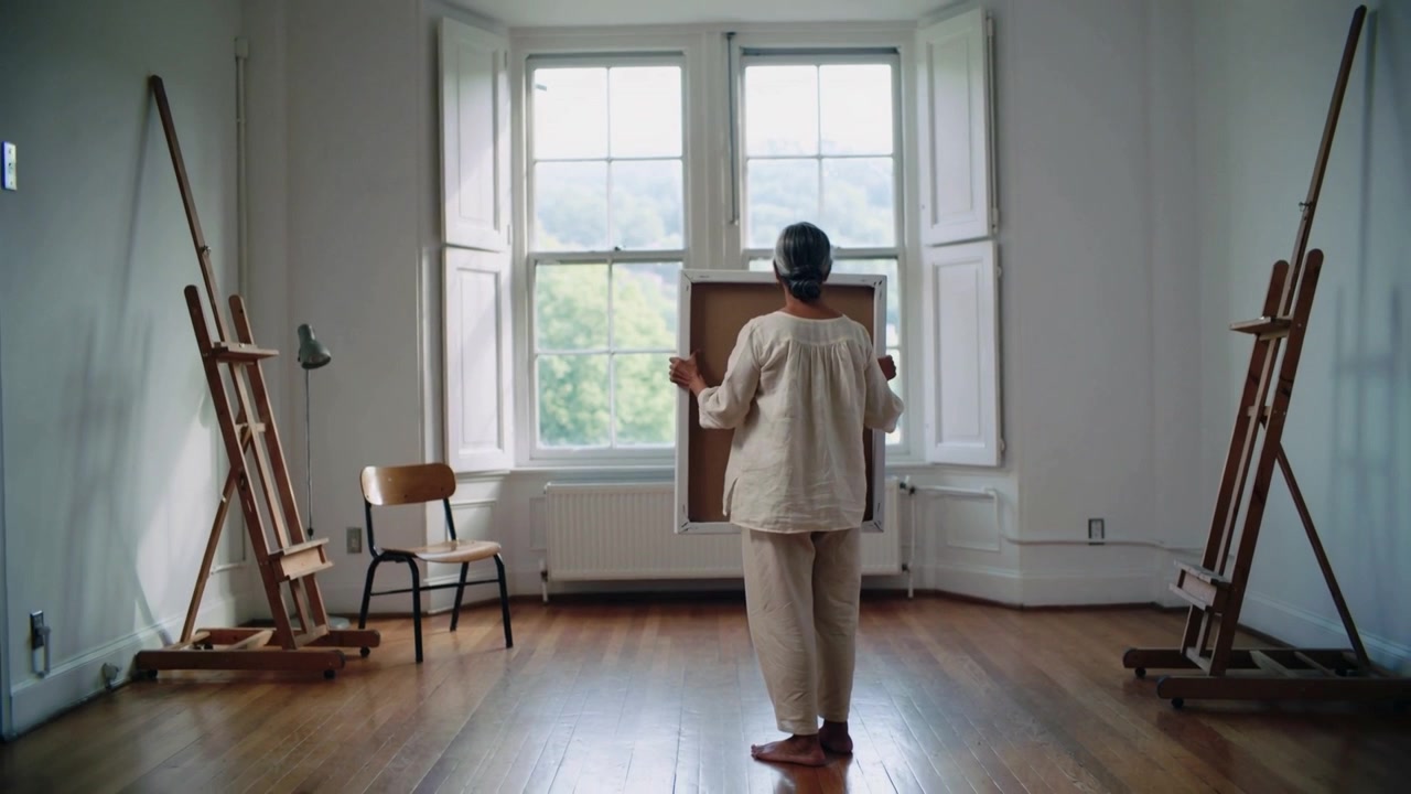 Holding canvas turning toward studio window Senior woman barefoot aligning with easels to place