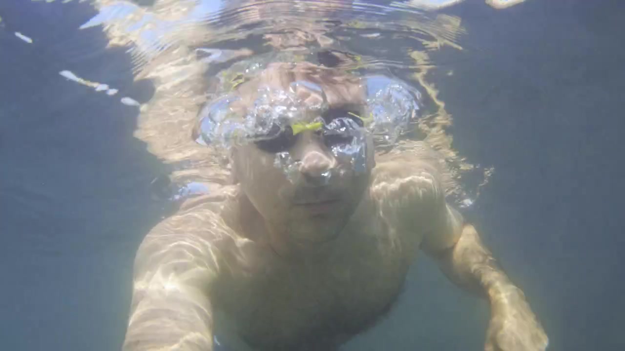 Hombre conteniendo la respiración bajo el agua con gafas de natación. Ondas de luz solar creando patrones en la piel. Ocio de verano y ejercicio acuático.
